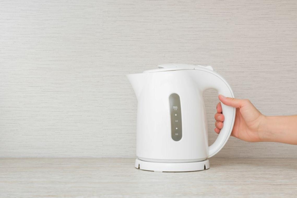 Young adult woman hand holding white plastic electric kettle on stone table top at home kitchen. Closeup. Front view.