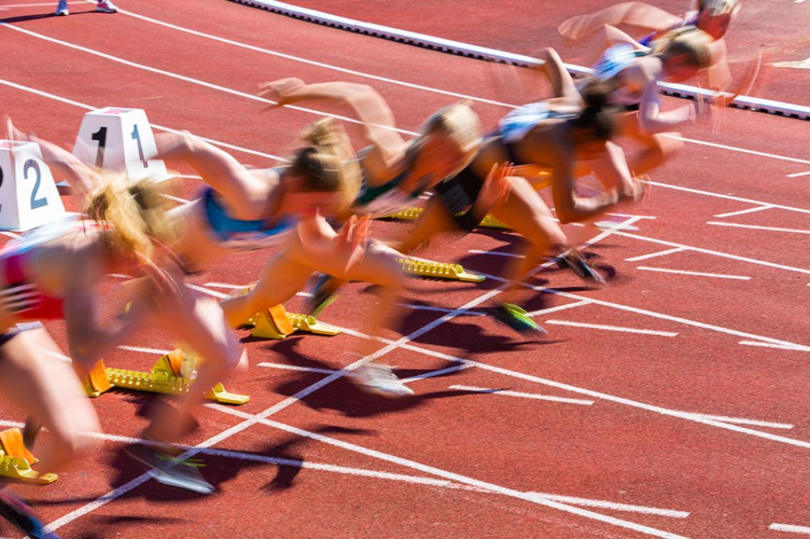 A blurred photo of female athletes at a starting block on a red athletics track. 
