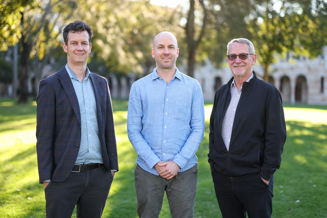 Three men stand in a grassy forecourt with sandstone buildings in the background.