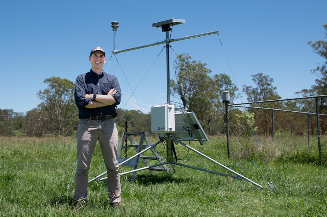 A man stands in a green paddock on a sunny day with a weather monitoring station behind him.