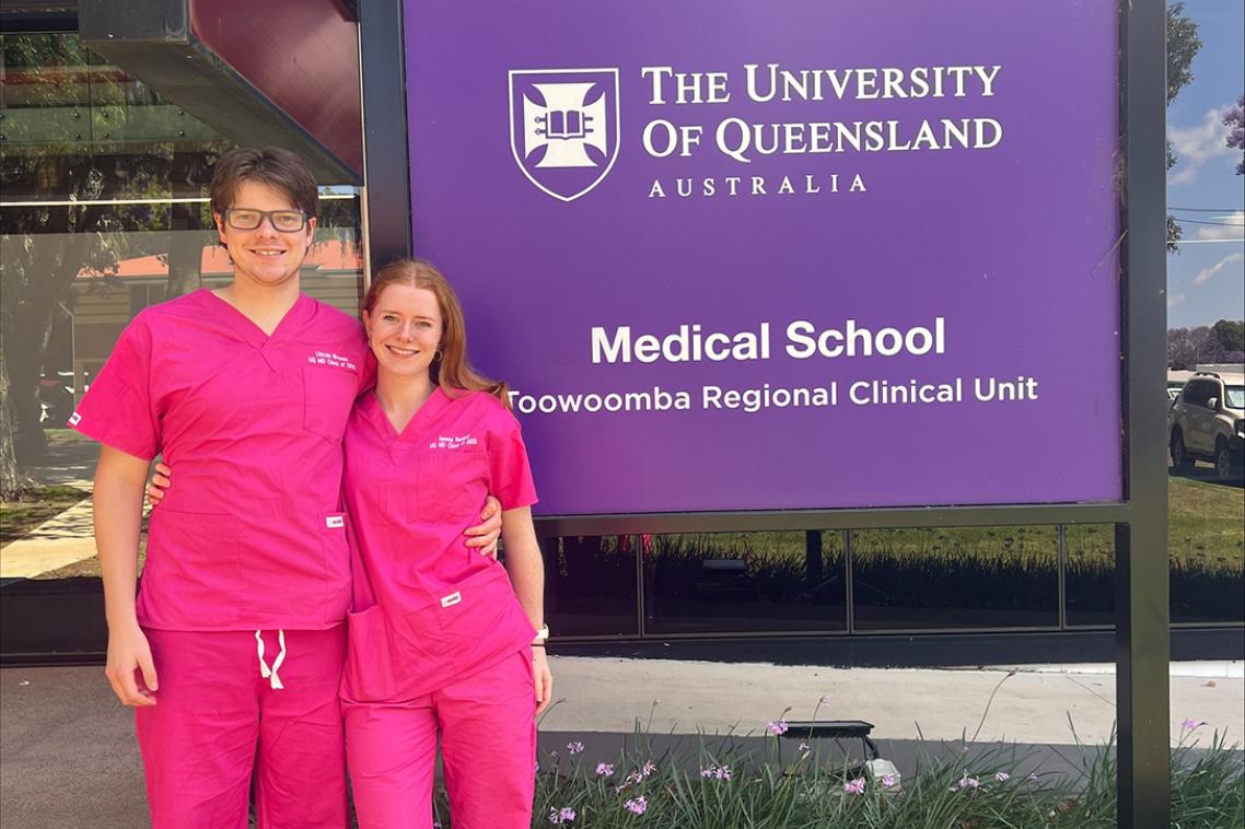A male and female student wearing pink scrubs taking a photo in front of a UQ sign 
