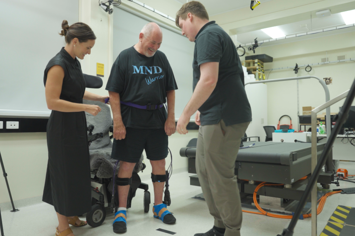 A man with a device fitted to his lower legs is in a lab setting with a man and woman either side of him.  the Neuromuscular Biomechanics Laboratory