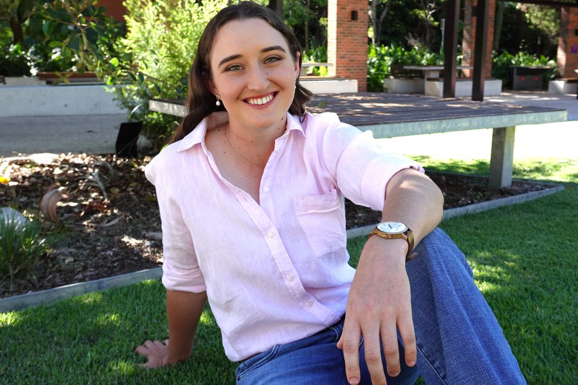 Piper sitting on the grass smiling at the camera while wearing a pink button up shirt and jeans.
