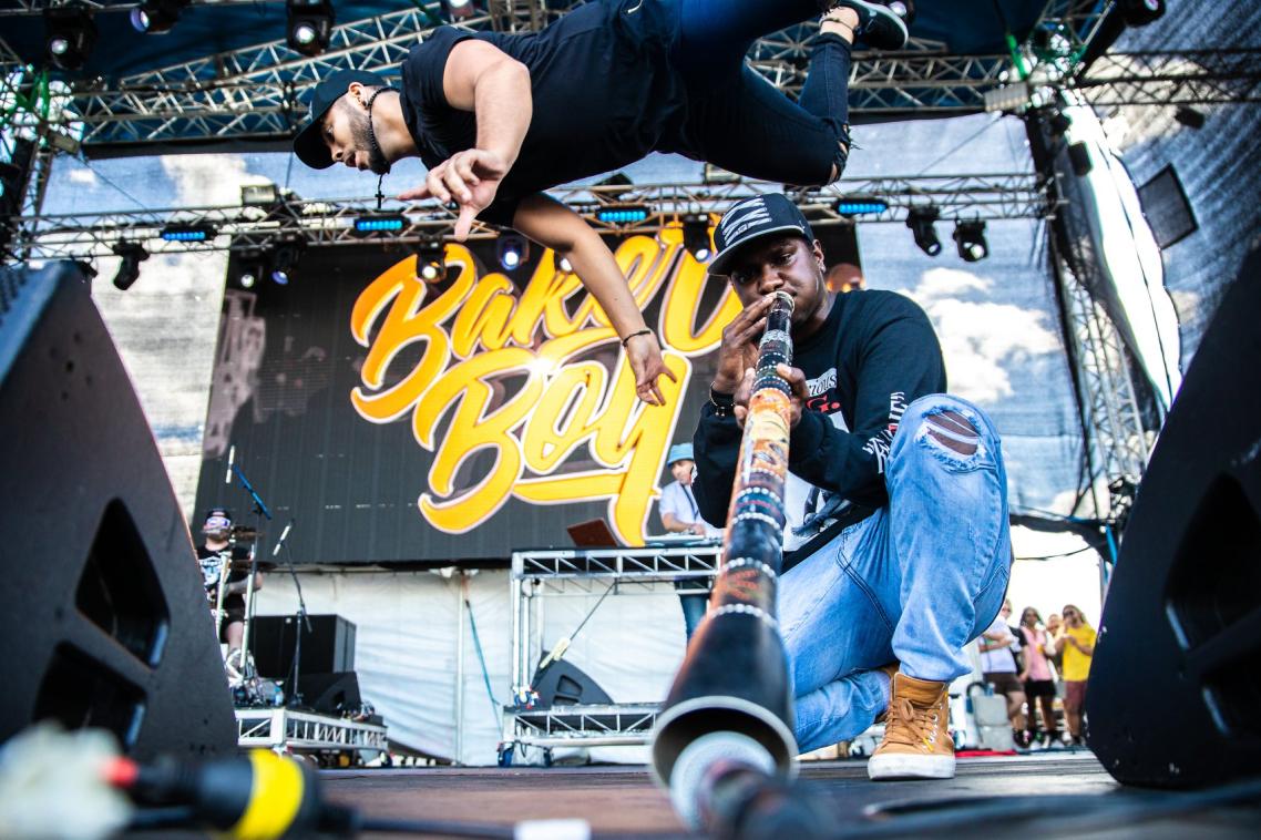 Baker Boy on stage. One performer is captured mid-flip, while another plays the didgeridoo, with the end of it pointing towards the camera.