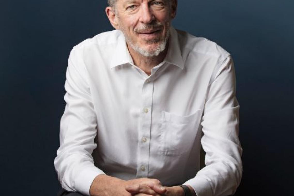 A man with grey hair and wearing a white shirt sits clasping his hands