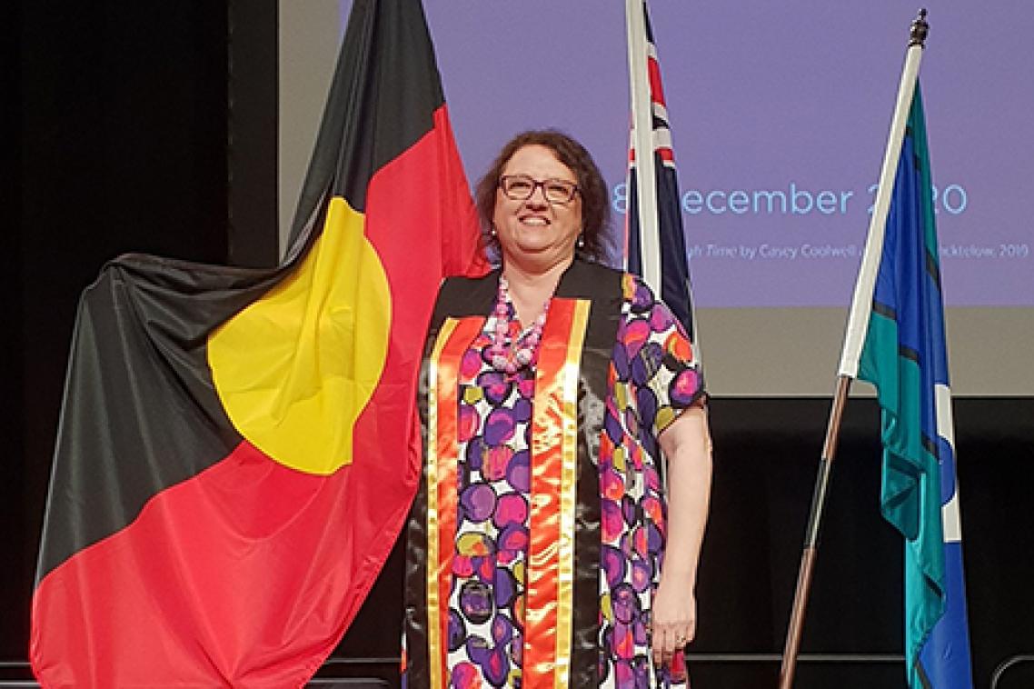 Lorelle Holland wearing a colourful dress and a black, red and yellow sash. She is standing in front of an Aboriginal, Australian, and Torres Strait Islands flag.
