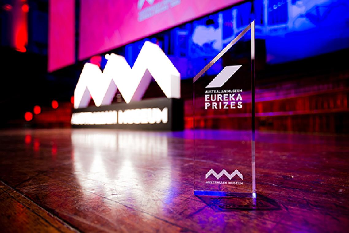 A glass trophy sitting on a wooden floor. Red and purple lights illuminate the room. There is a white sign reading Australian Museum in the background.