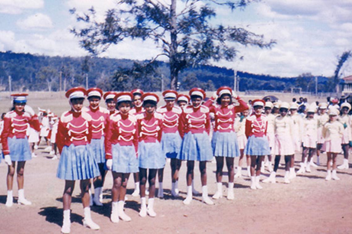 A group of teenage girls next to a smaller group of young girls. The teenagers are wearing pale blue short pleated skirts, red hats, red shirts with tassels, mid-length white socks, and white shoes. The younger children are wearing white dresses, hats, socks and shoes.