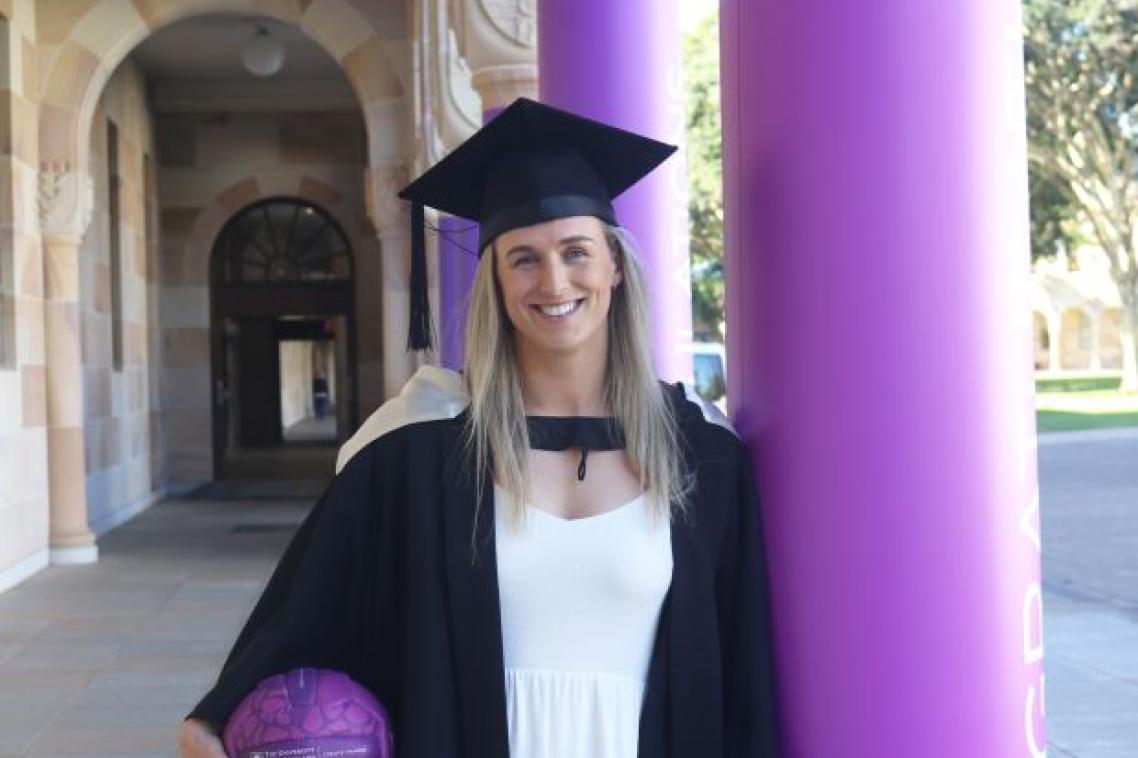 A young woman in a graduation cap and gown holds a netball and leans against a column in the St Lucia Great Court