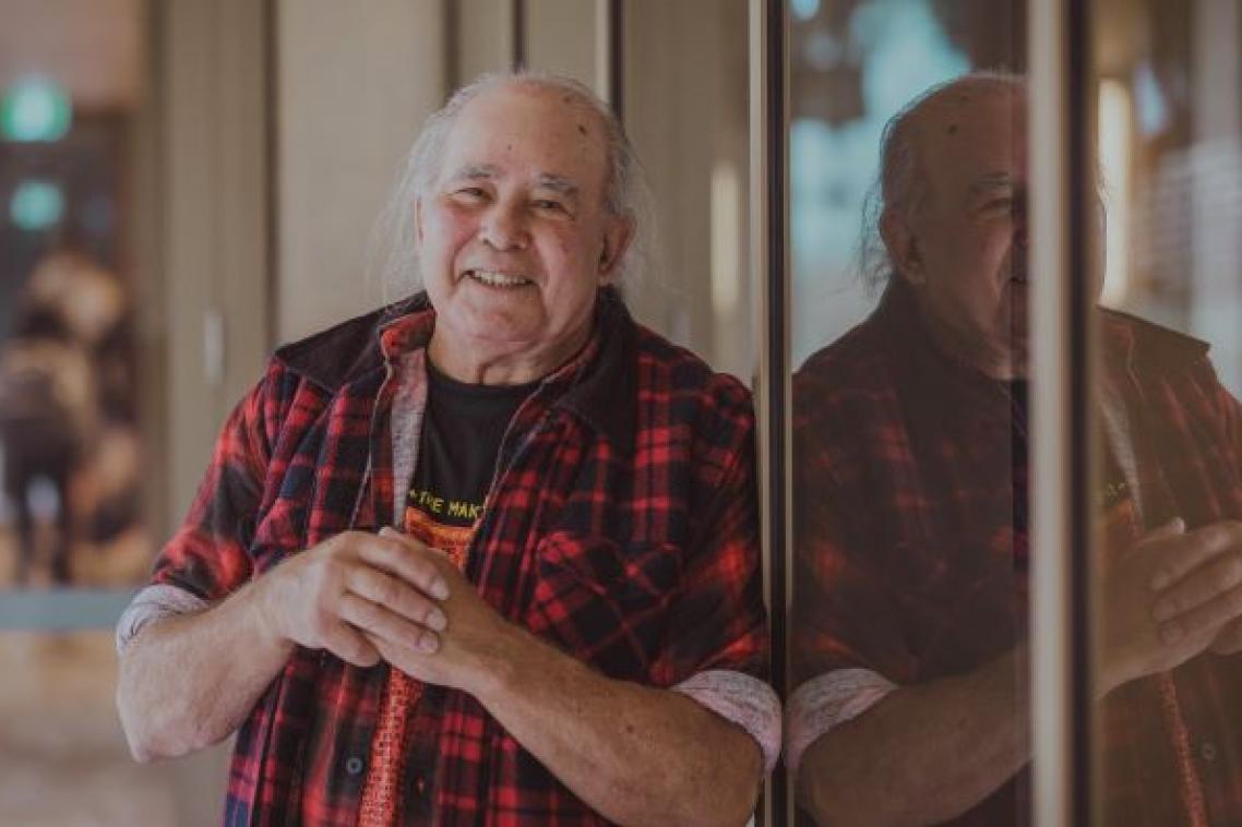 a man in a check shirt leans against a window in which he is reflected