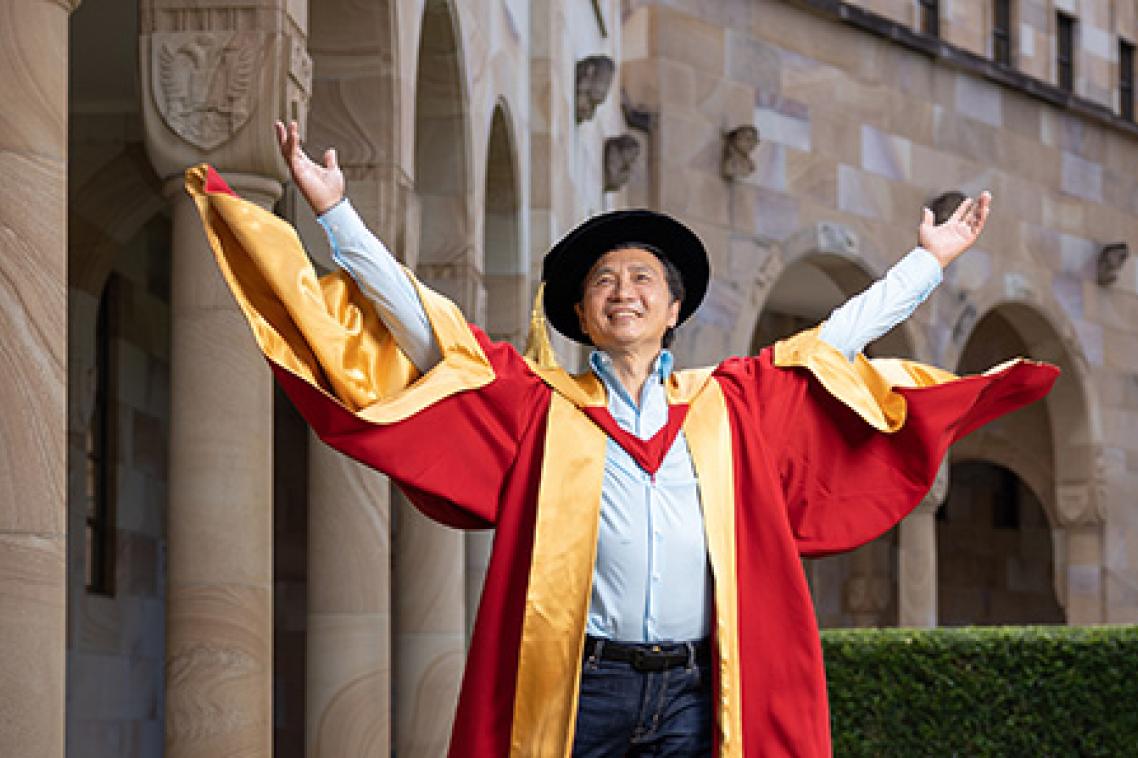 Li Cunxin AO stands in the Great Court wearing his red and yellow academic dress and cap.
