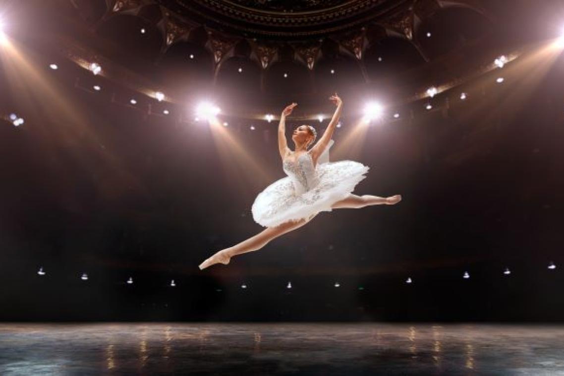 A ballet dancer in white tutu leaping under lights on stage. Adobe