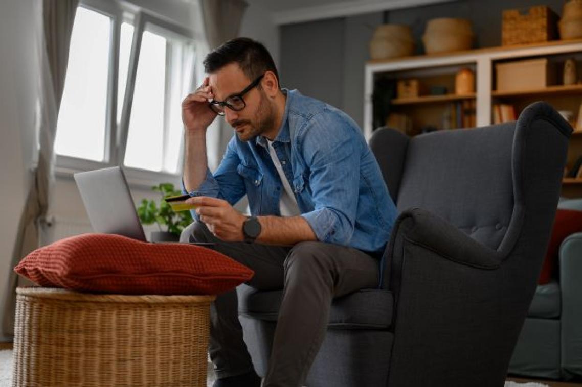 Man sitting at laptop with credit card in hand looking worried. Adobe.
