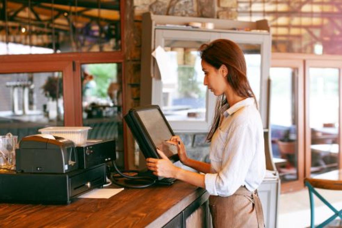 A hospitality worker stands at the counter of a cafe, typing into a computer.