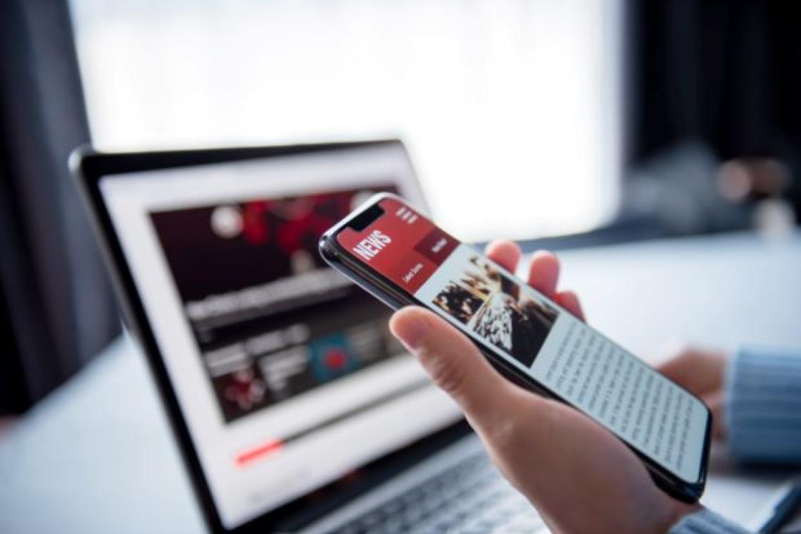 hand holding a phone with screen on a news site in front of a laptop on a news web page