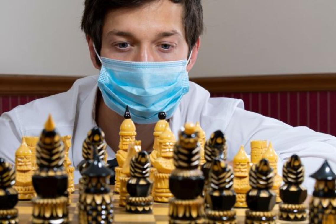 A close up of the face of a man wearing a surgical mask behind a chess board set with playing pieces