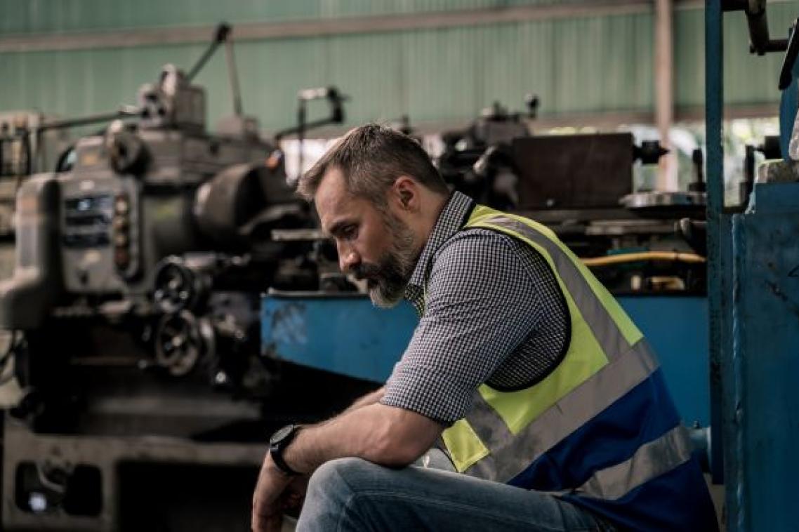 A bearded man in a short and high vis vest sits with his head bowed in a factory.