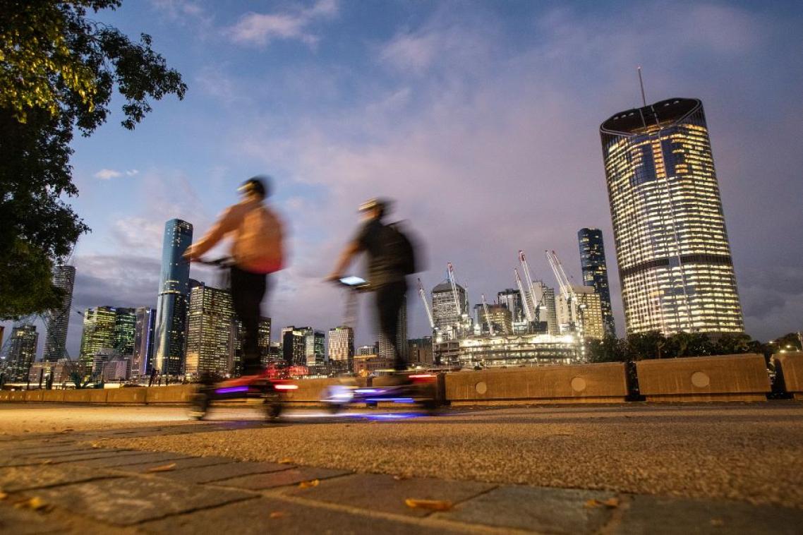 two people on scooters riding along a footpath at dusk. The city skyline is in the background