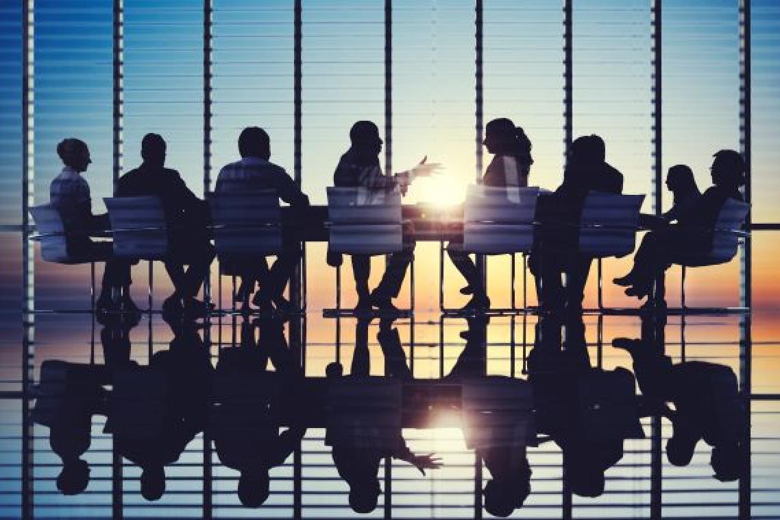 Men and women sitting around a board room table with sun shining through glass wall. Adobe
