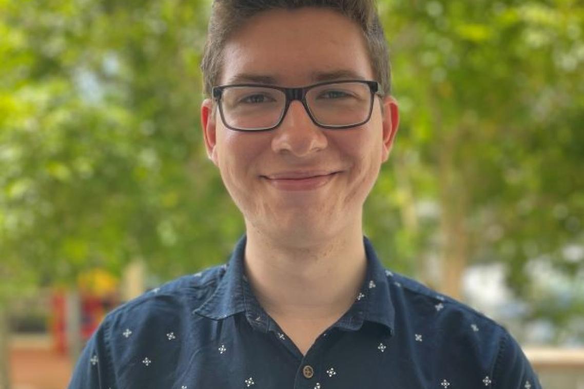A young man in glasses and a navy button-up shirt smiles at the camera. There is a leafy green tree in the background.