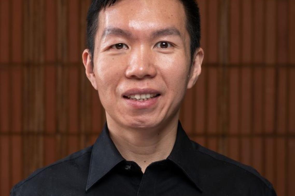 A man with black hair and wearing a black collared shirt stands in front of a brown timber wall. He is smiling at the camera.