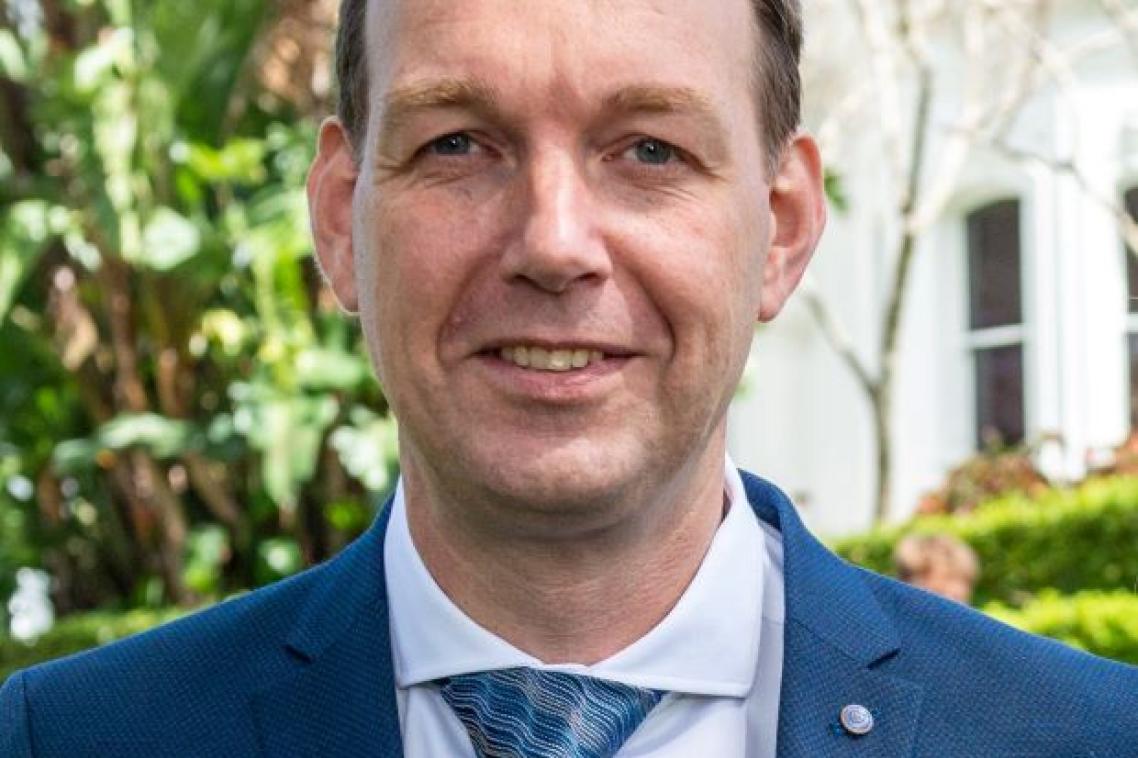 A man in a tie, white shirt and blue jacket smiles. He is outdoors with a building and tree in the background.