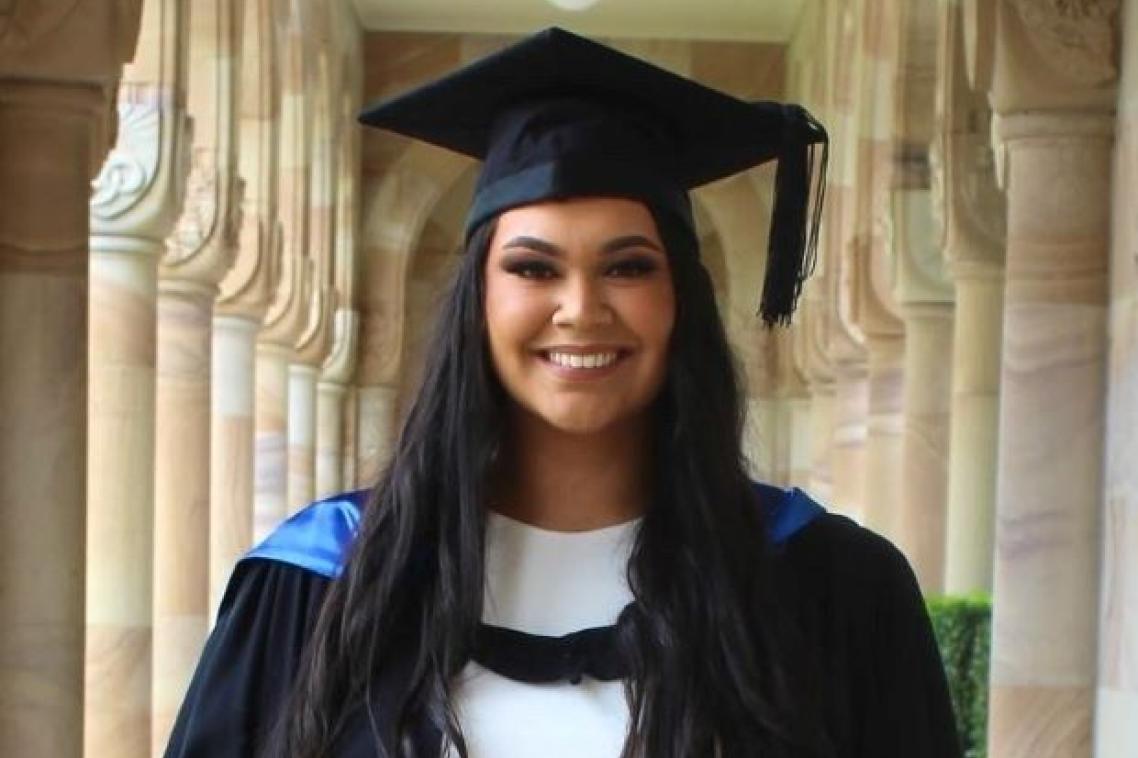 A woman with long black hair is smiling at the camera, she is wearing a graduation cap and gown over a white dress and is standing in the sandstone cloisters of UQ's Great Court.