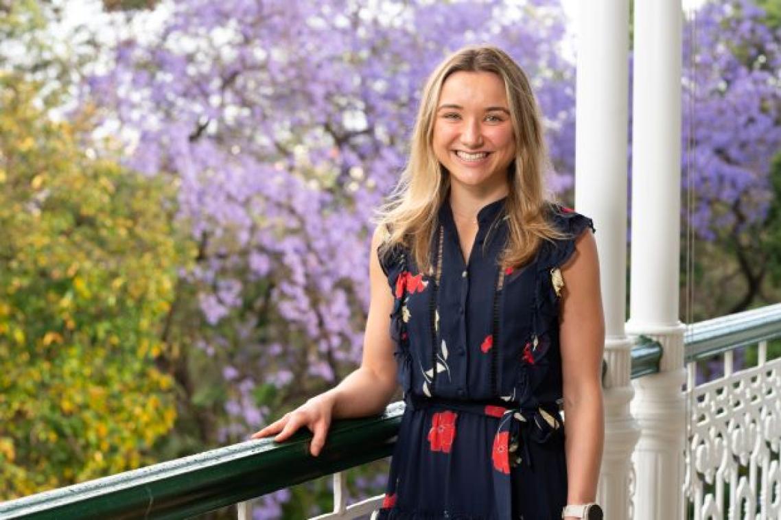 A young woman wearing a navy floral dress stands holding a railing with a flowering jacaranda tree in the background. She has blonde hair and is smiling.