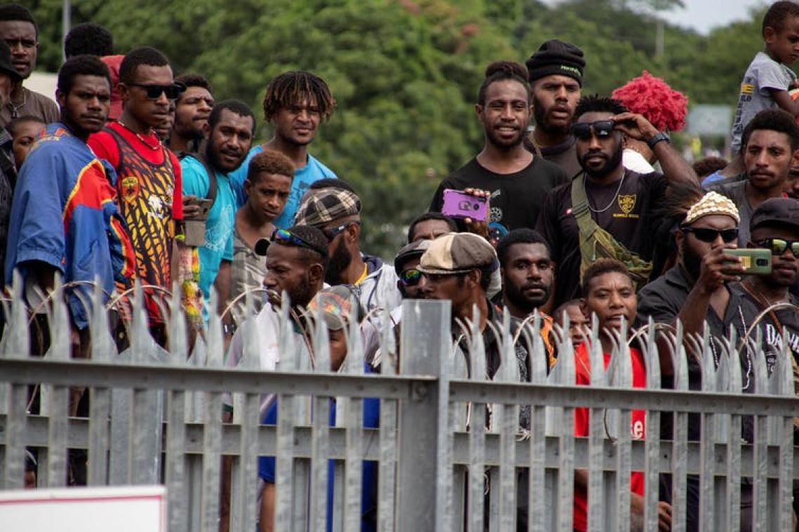 A group of about 16 men from PNG stand watching something over a fence, some with cameras