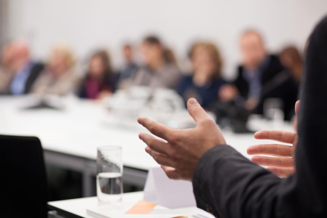 A business-man's hands gesture. In the background are people gathered around a conference