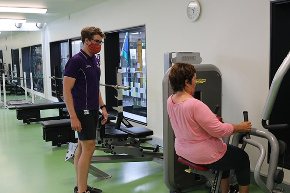 Woman using lat pulldown machine under supervision of man in UQ shirt