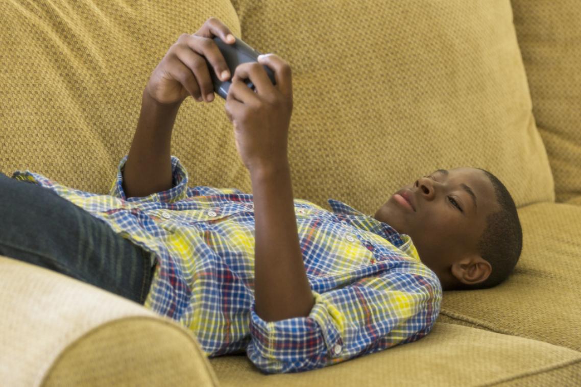 A boy lays on a couch watching something on a hand-held screen device.