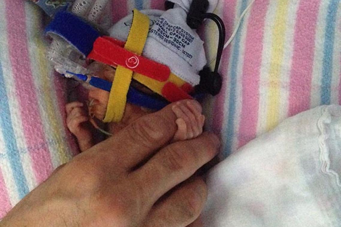 Hand holding the tiny hand of a premature baby, which is wearing a beanie and attached to tubes
