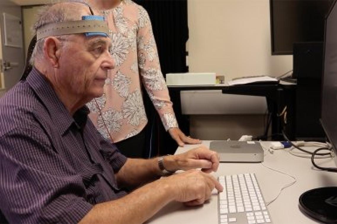 Man sitting at computer with electrical stimulation machine on head with researcher standing over his shoulder.