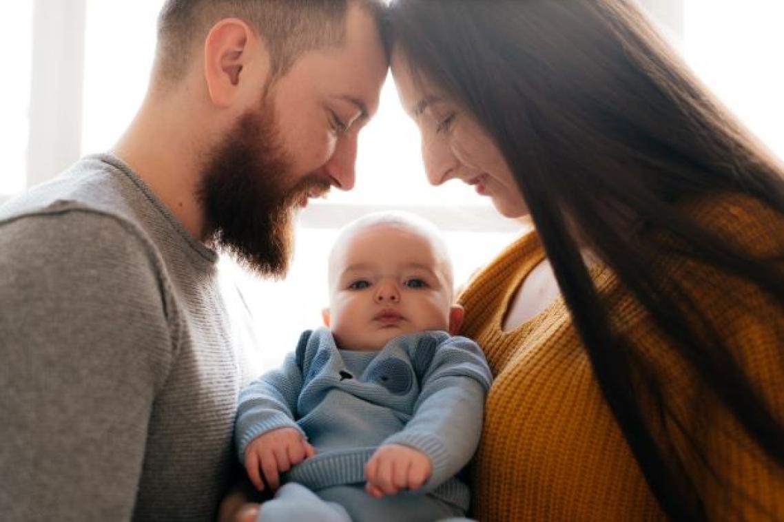a man with a beard and a woman with long dark hair look down at a baby wearing blue between them