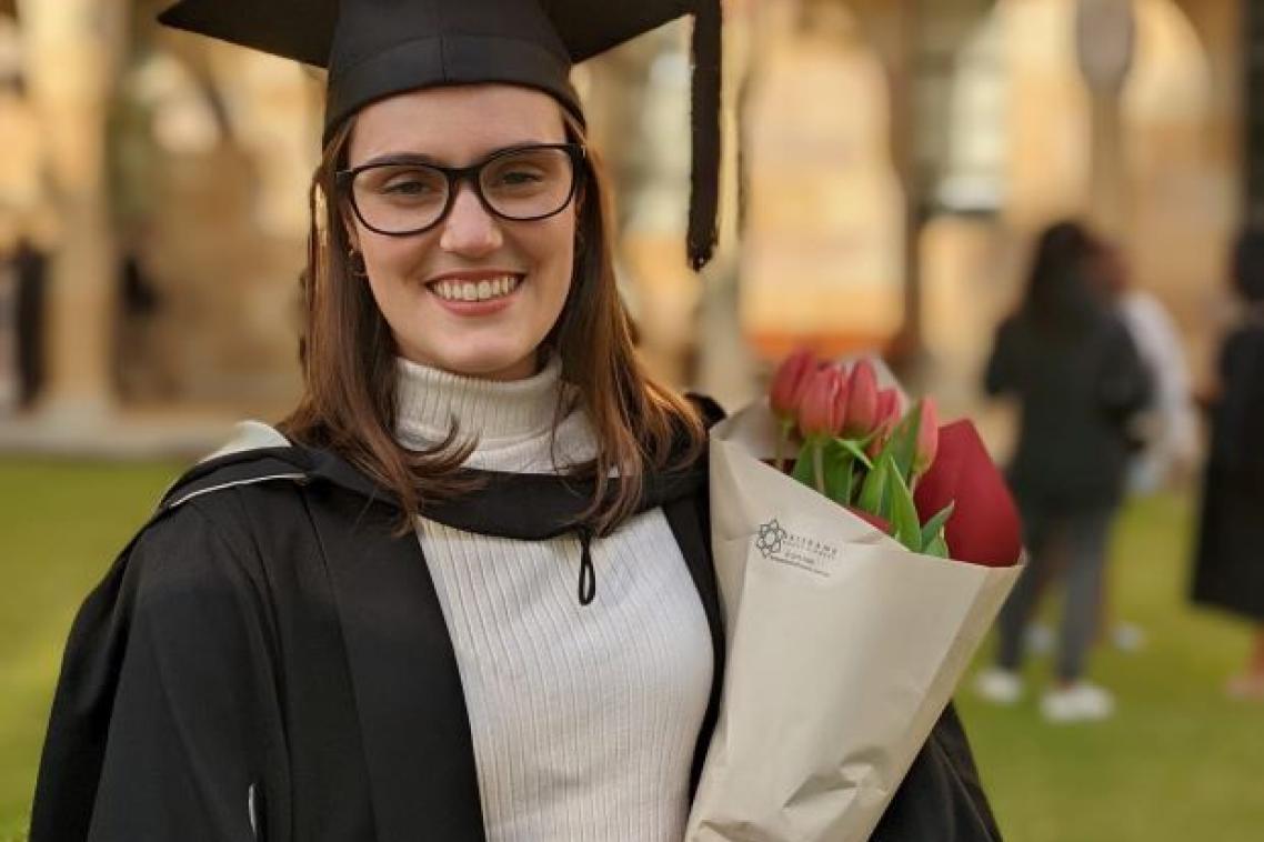 A young woman faces the camera wearing graduation robe and cap, smiling. She is holding a bunch of flowers.