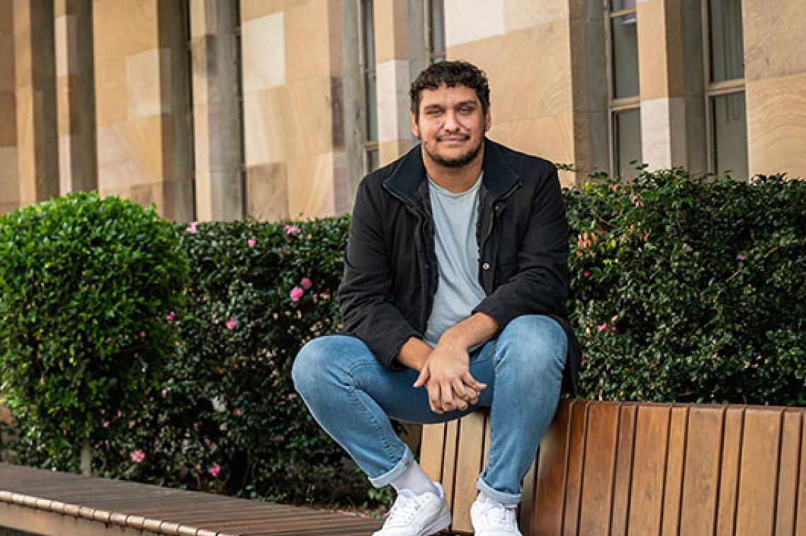 A man sits on the back of an outdoor bench with a garden and sandstone building in the background.