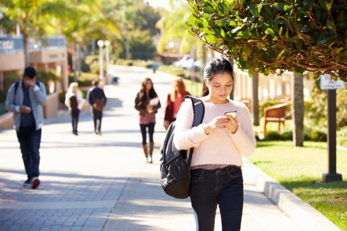 A street in daylight with people with pedestrians looking at their phones as they walk with a young woman in the foreground.