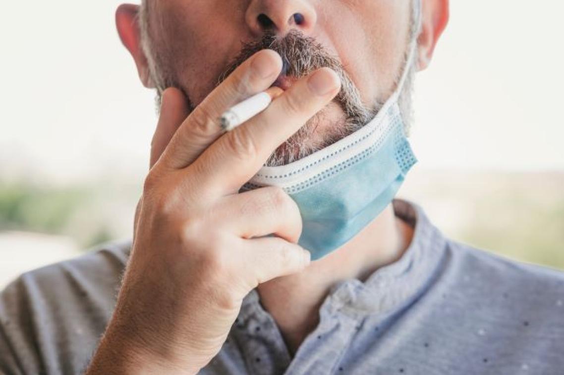 A close up of a man wearing a disposable mask over his chin, holding a cigarette to his lips.