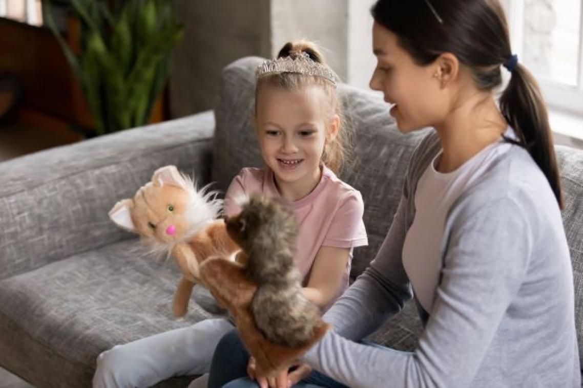 a child and a woman sit on a grey couch holding two animal puppets