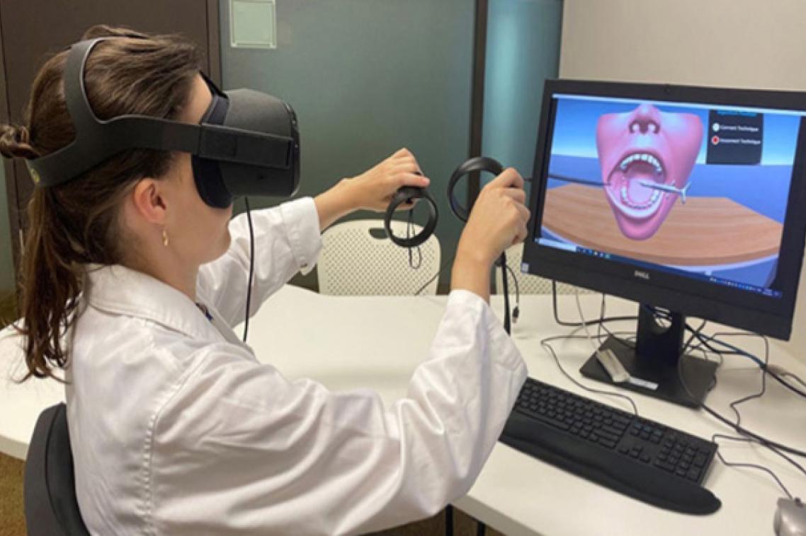 a person in white coat wears virtual reality glasses and holds handsets in front of a screen with an image of a mouth and teeth