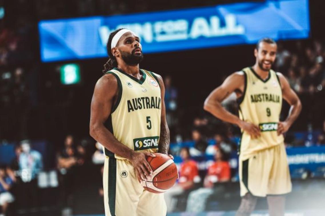 An Indigenous basketball player wearing an Australian uniform holds a basketball and prepares to shoot. There is a teammate and a stadium crowd in the background.