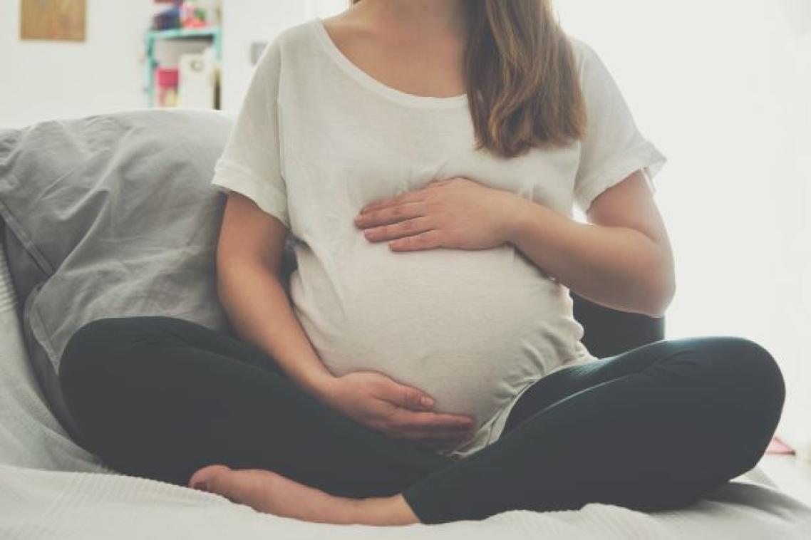 A heavily pregnant woman sits cross-legged with her arms protectively around the top and bottom of her belly.