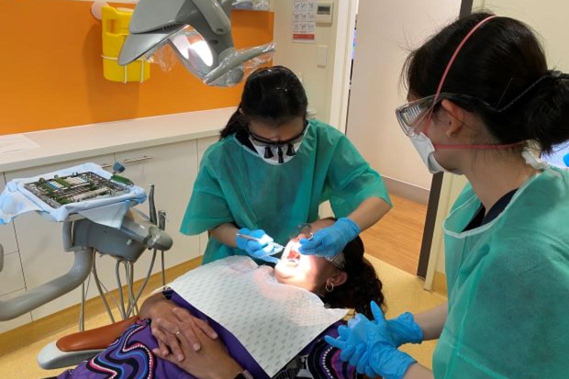 A patient lies on a reclining chair in a dental clinic as two student dentists in scrubs treat her.