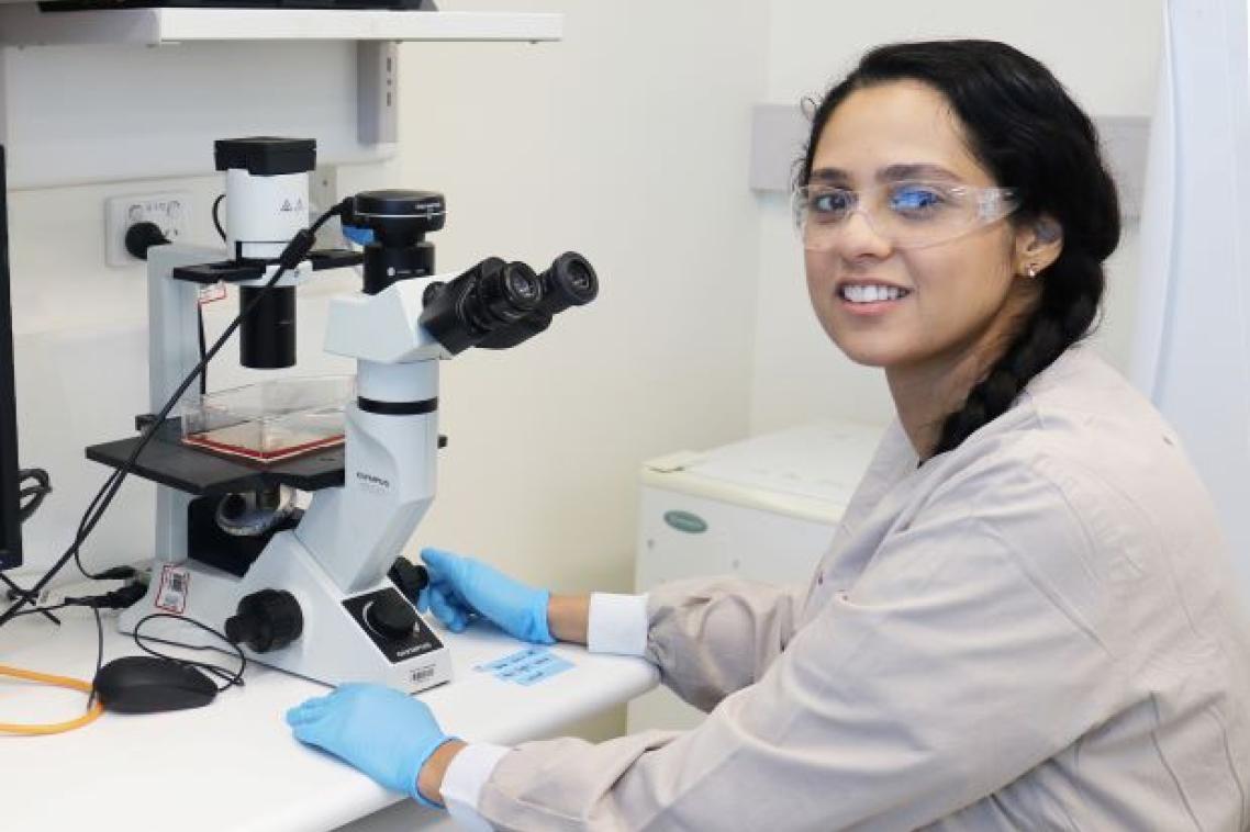 a woman with dark hair wearing safety glasses sits in front of a white and black microscope