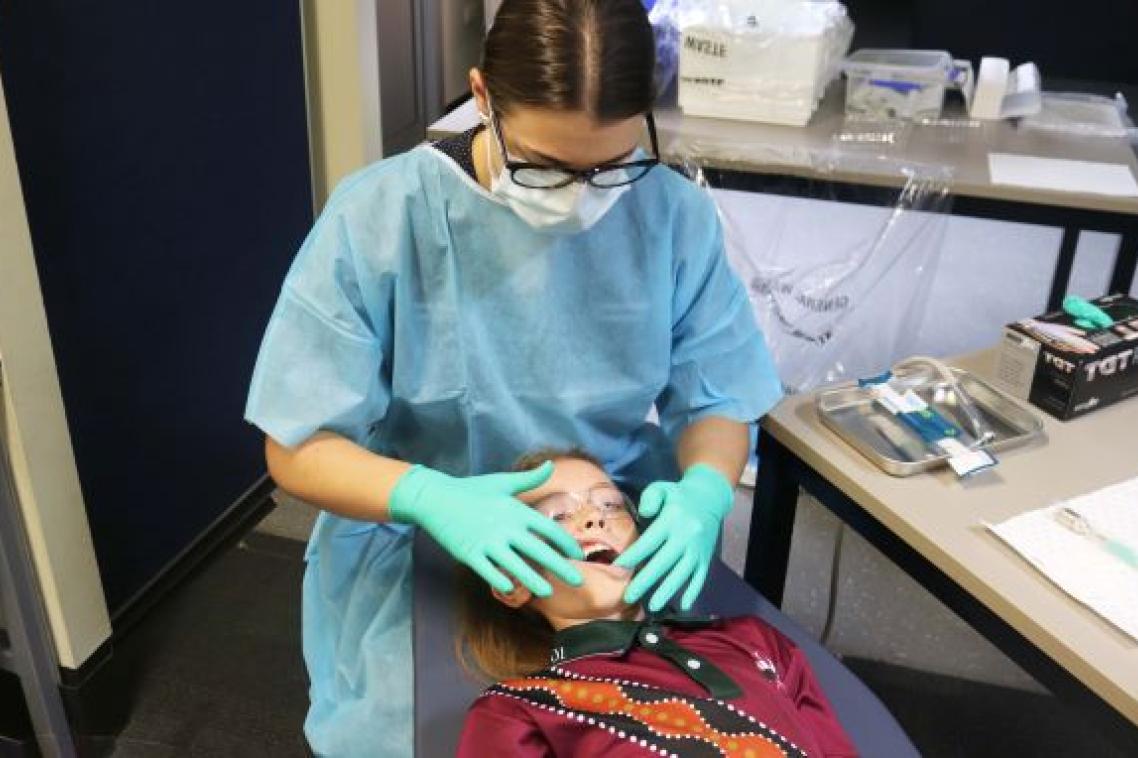 A woman in a blue gown, green gloves and facemask looks at the teeth of a child lying in front of her