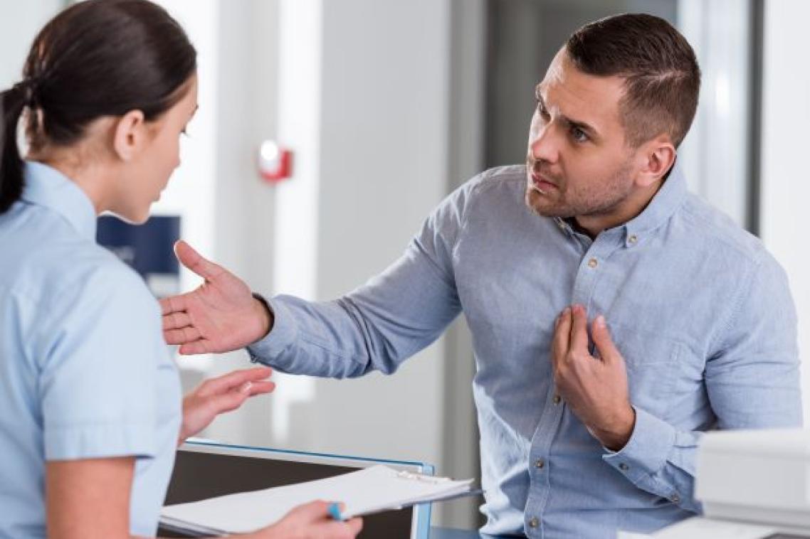 A man looking aggressive and threatening stands before a woman at a medical reception desk.