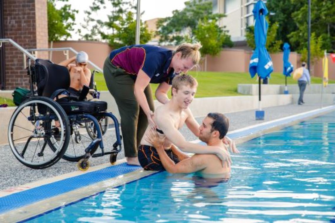 A young man is being helped from a wheelchair into a swimming pool by two trainers.
