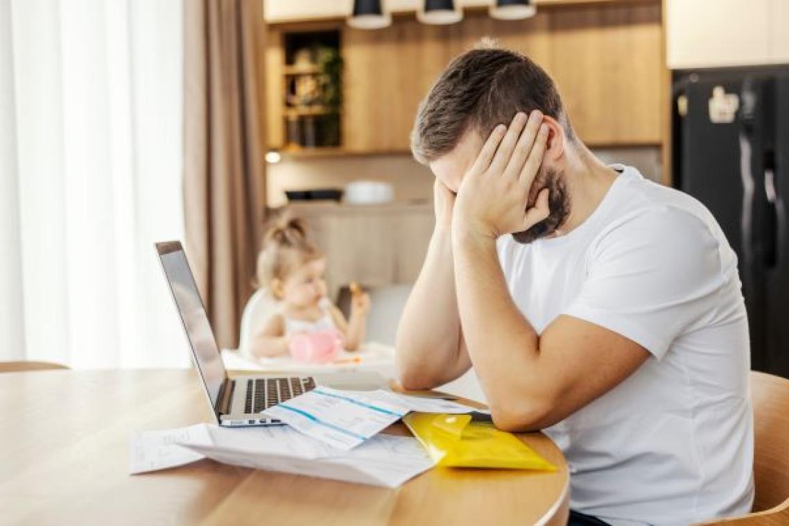 a man sits at a table with his face in his hands, in front of him are papers and a laptop, behind him is a small child in a high chair