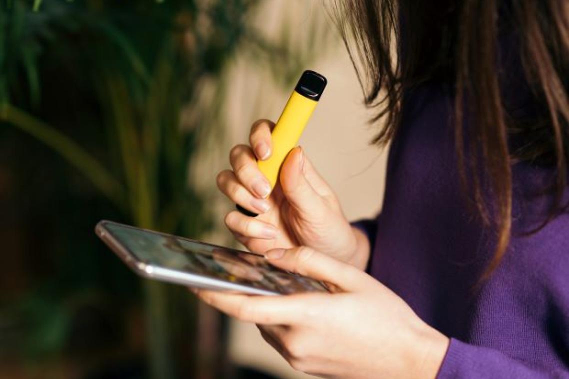 A young woman in profile with long dark hair falling across her face, holding a vape and a mobile phone.